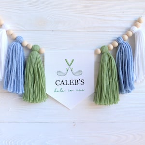 May include: A decorative banner with white, blue, and green yarn tassels and wooden beads. The banner features a white pennant with the text "CALEB'S hole in one" and a watercolor illustration of crossed golf clubs.