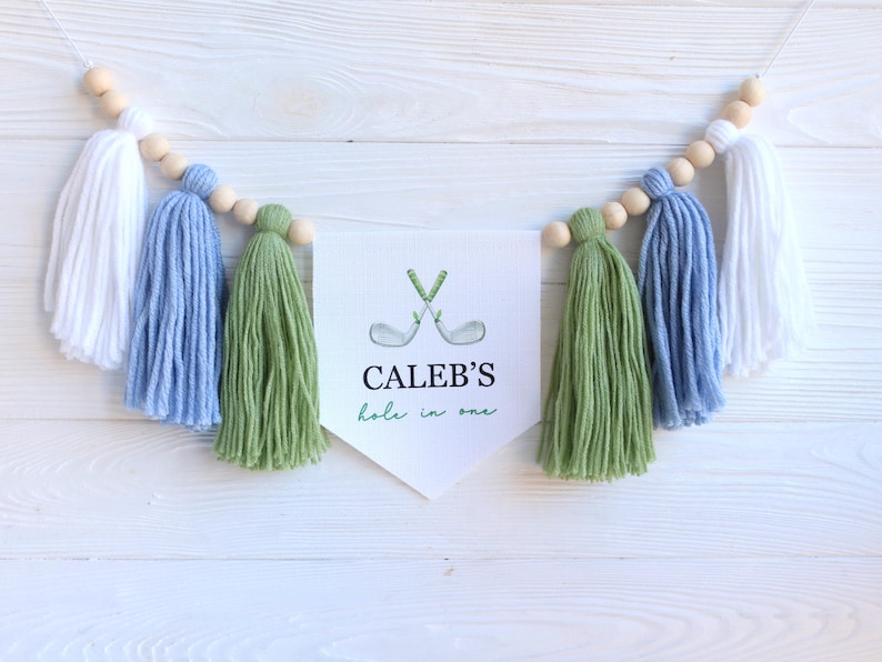 May include: A decorative banner with white, blue, and green yarn tassels and wooden beads. The banner features a white pennant with the text "CALEB'S hole in one" and a watercolor illustration of crossed golf clubs.