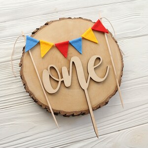 May include: A wooden cake topper with the word "one" in cursive script, surrounded by a string of colorful felt flags. The flags are red, yellow, and blue.