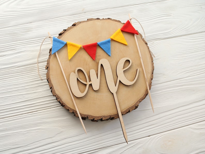 May include: A wooden cake topper with the word "one" in cursive script, surrounded by a string of colorful felt flags. The flags are red, yellow, and blue.