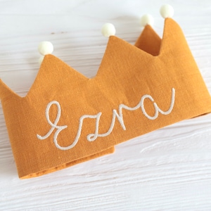 May include: A yellow linen crown with white pom-poms around the edge and the name "Ezra" embroidered in white thread.