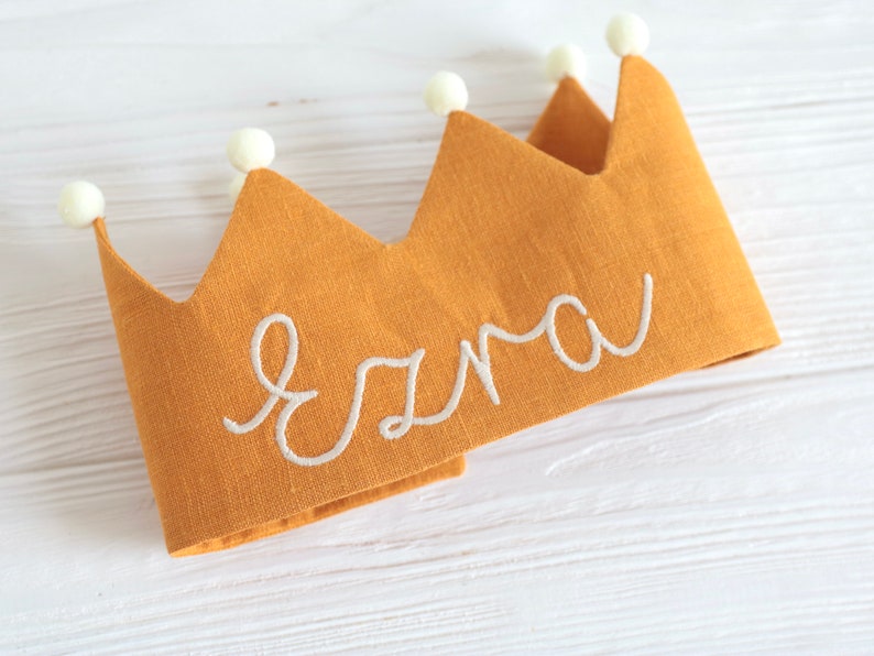 May include: A yellow linen crown with white pom-poms around the edge and the name "Ezra" embroidered in white thread.