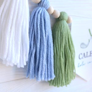May include: A handmade tassel garland featuring white, light blue, and green yarn tassels strung on a wooden bead garland. A white card with the text "CALEB'S" and golf-themed graphics is visible.
