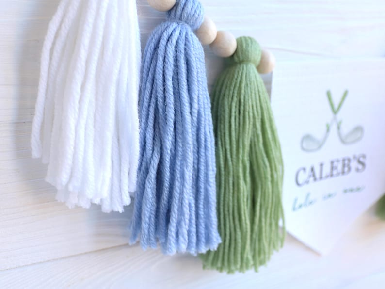 May include: A handmade tassel garland featuring white, light blue, and green yarn tassels strung on a wooden bead garland. A white card with the text "CALEB'S" and golf-themed graphics is visible.