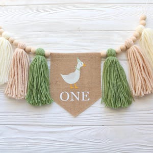 May include: A burlap banner with a white duck illustration and the word "ONE" in white letters. The banner is strung with wooden beads and yarn tassels in white, peach, and green.