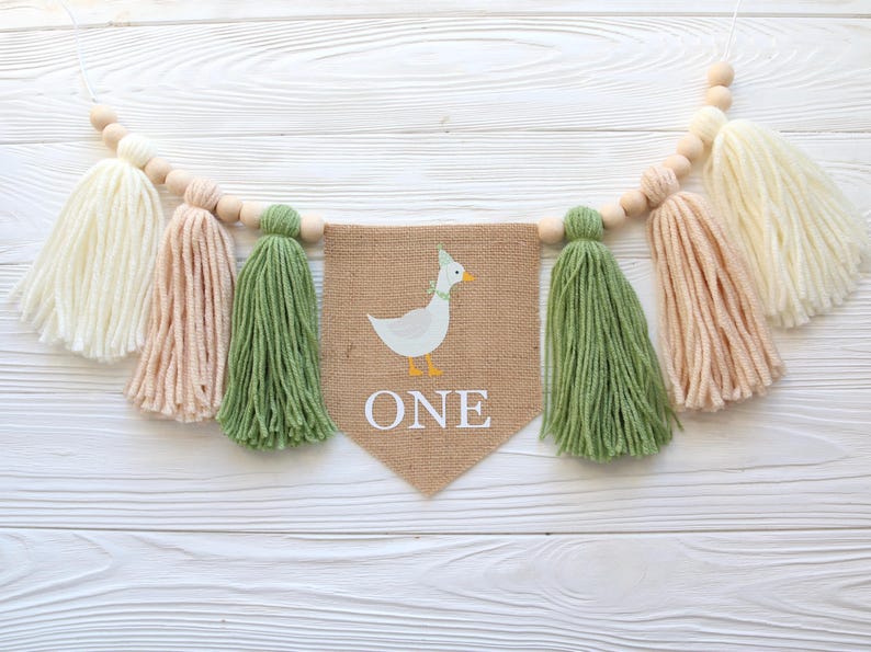 May include: A burlap banner with a white duck illustration and the word "ONE" in white letters. The banner is strung with wooden beads and yarn tassels in white, peach, and green.