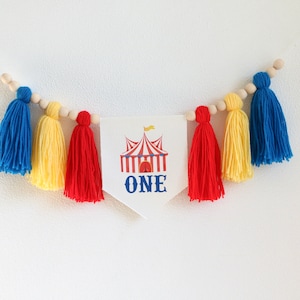 May include: A white banner with a red and white striped circus tent and the word "ONE" in blue. The banner is strung with wooden beads and yellow, red, and blue yarn tassels.