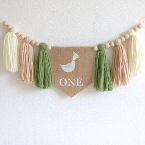 May include: A burlap banner with a white duck and the word "ONE" in white letters. The banner is strung with wooden beads and green and beige yarn tassels.