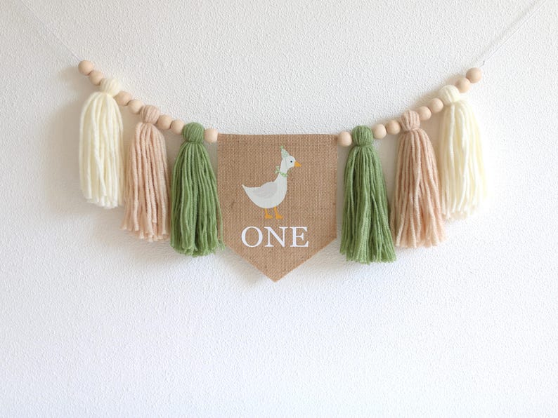 May include: A burlap banner with a white duck and the word "ONE" in white letters. The banner is strung with wooden beads and green and beige yarn tassels.