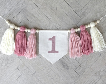 May include: A white banner with the number "1" in pink, decorated with pink and white yarn tassels and wooden beads. The banner is strung on a white cord.