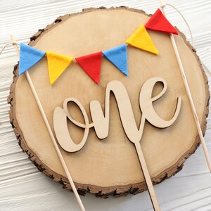 May include: A wooden cake topper with the word "one" in cursive script. The topper is decorated with a string of felt flags in red, yellow, and blue.