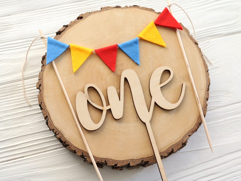 May include: A wooden cake topper with the word "one" in cursive script. The topper is decorated with a string of felt flags in red, yellow, and blue.