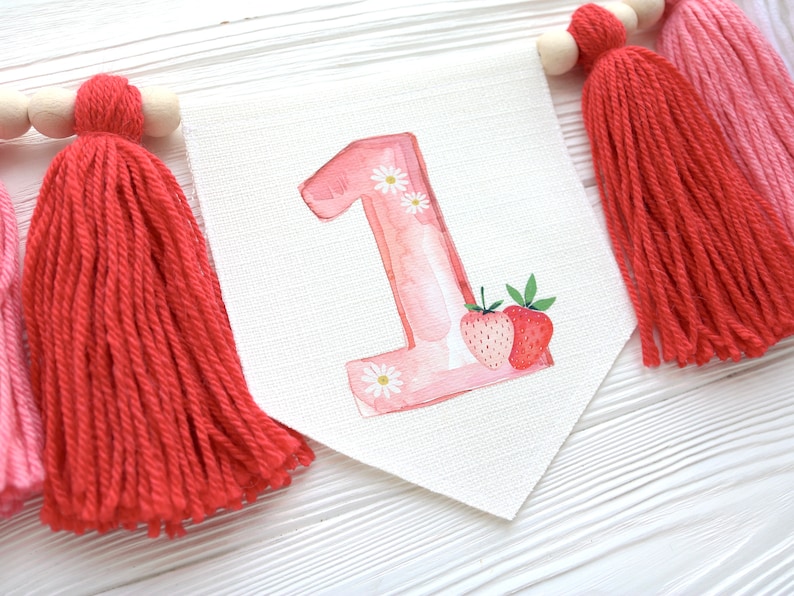 Puede incluir: Un n&uacute;mero uno en acuarela rosa y blanco con flores blancas y dos fresas rojas. El n&uacute;mero est&aacute; unido a una pancarta blanca con borlas rojas y rosas.