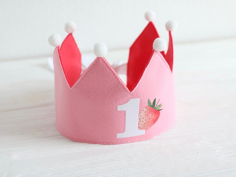 Puede incluir: Corona de fieltro rosa con pompones blancos y un n&uacute;mero uno de fieltro blanco y un dise&ntilde;o de fresa.