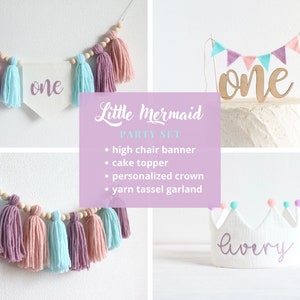May include: A Little Mermaid themed first birthday party set with a pink and blue yarn tassel garland, a cake topper with a wooden "one" and a pink and blue bunting, and a white crown with the name "lavery" embroidered in purple.