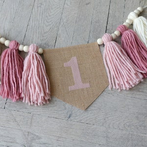 May include: A burlap banner with the number "1" printed in pink, decorated with a string of wooden beads and pink and white yarn tassels.