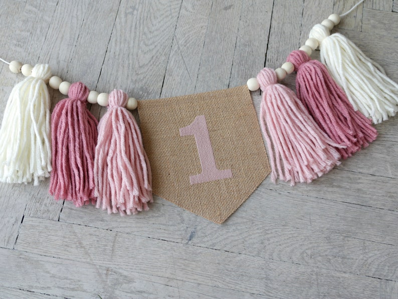 May include: A burlap banner with the number "1" printed in pink, decorated with a string of wooden beads and pink and white yarn tassels.