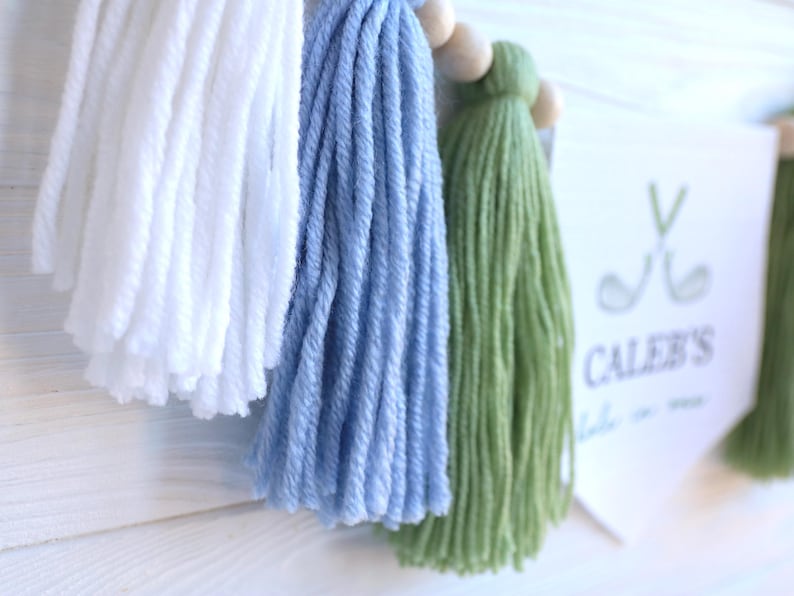 May include: A handmade banner with white, light blue, and green yarn tassels. The banner includes wooden beads and a white pennant with the text "Caleb's" and golf-themed graphics.