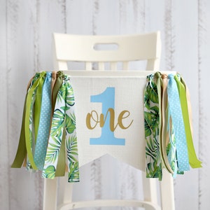Puede incluir: Una pancarta de silla alta blanca con un n&uacute;mero uno azul y la palabra "one" en letras doradas. La pancarta tiene tela de hojas tropicales verdes y blancas a cada lado y cintas azules, verdes y marrones que cuelgan de la parte inferior.