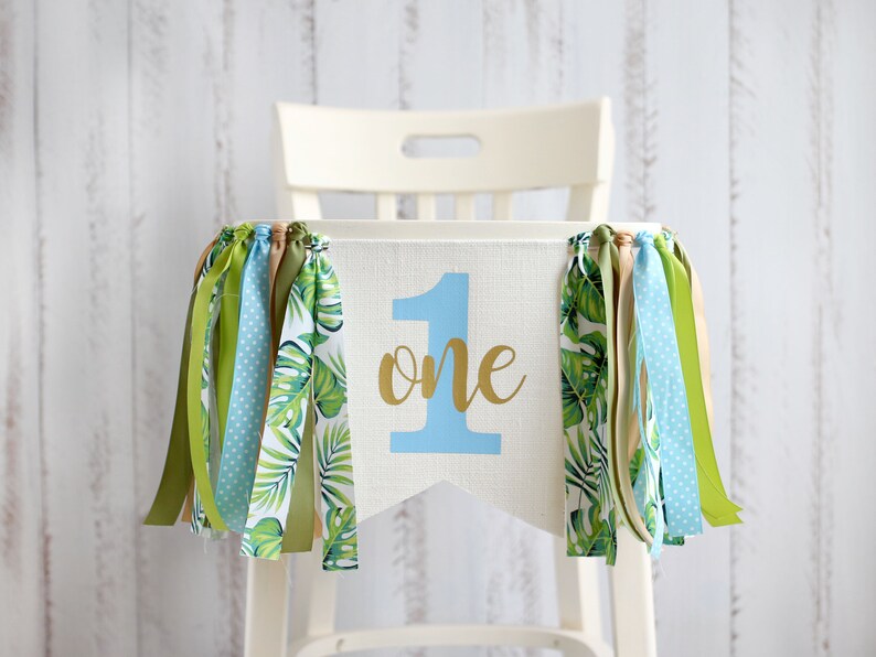 Puede incluir: Una pancarta de silla alta blanca con un n&uacute;mero uno azul y la palabra "one" en letras doradas. La pancarta tiene tela de hojas tropicales verdes y blancas a cada lado y cintas azules, verdes y marrones que cuelgan de la parte inferior.