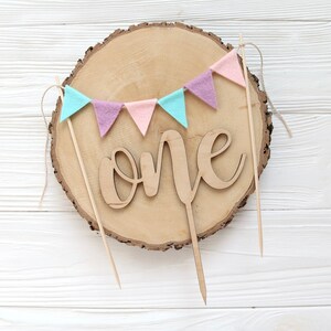 May include: A wooden cake topper with the word "one" in cursive script, surrounded by a garland of pastel pink, purple, and blue felt flags. The topper is on a wooden slice.