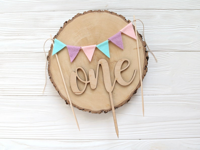 May include: A wooden cake topper with the word "one" in cursive script, surrounded by a garland of pastel pink, purple, and blue felt flags. The topper is on a wooden slice.