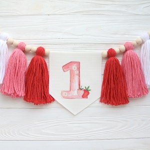 Puede incluir: Una pancarta rosa y blanca con el n&uacute;mero "1" en rosa con flores blancas y un dise&ntilde;o de fresa. La pancarta est&aacute; enhebrada con borlas blancas, rosas y rojas.
