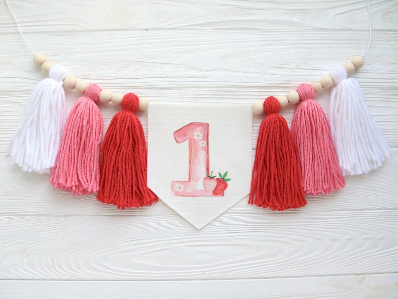 Puede incluir: Una pancarta rosa y blanca con el n&uacute;mero "1" en rosa con flores blancas y un dise&ntilde;o de fresa. La pancarta est&aacute; enhebrada con borlas blancas, rosas y rojas.