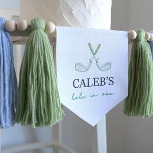 May include: A banner with the text "CALEB'S hole in one" and crossed golf clubs. The banner is flanked by blue and green tassels, with wooden beads. The banner is likely for a golf-themed celebration.