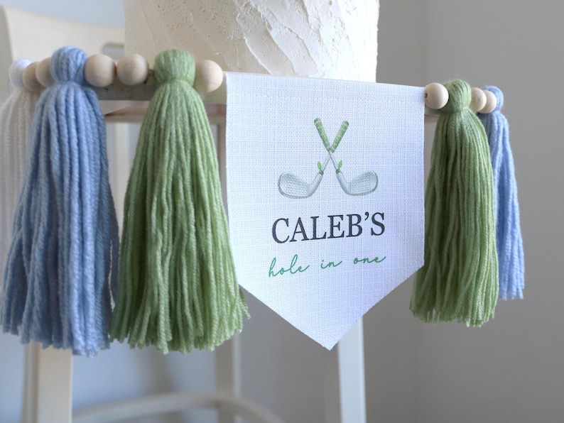 May include: A banner with the text "CALEB'S hole in one" and crossed golf clubs. The banner is flanked by blue and green tassels, with wooden beads. The banner is likely for a golf-themed celebration.