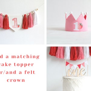 Puede incluir: Una corona de fieltro rosa con pompones blancos y un n&uacute;mero uno blanco con un dise&ntilde;o de fresa. Un pastel blanco con una guirnalda de borlas rosa y blanca y un adorno de pastel de madera que dice "one" en cursiva.