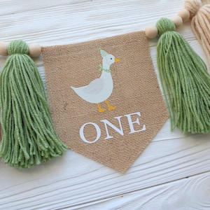 May include: A burlap banner with a white duck wearing a party hat and the word "ONE" in white letters. The banner is decorated with green and cream tassels.