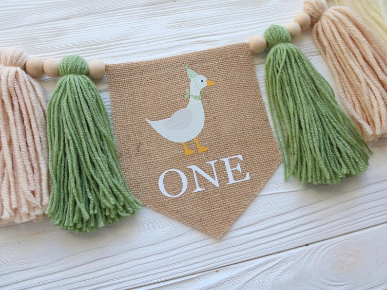 May include: A burlap banner with a white duck wearing a party hat and the word "ONE" in white letters. The banner is decorated with green and cream tassels.