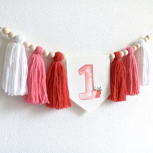 Puede incluir: Una pancarta blanca con el n&uacute;mero "1" en acuarela rosa con un dise&ntilde;o de fresa. La pancarta est&aacute; enhebrada con cuentas de madera y borlas rosas y rojas.