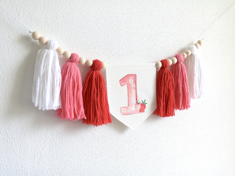 Puede incluir: Una pancarta blanca con el n&uacute;mero "1" en acuarela rosa con un dise&ntilde;o de fresa. La pancarta est&aacute; enhebrada con cuentas de madera y borlas rosas y rojas.