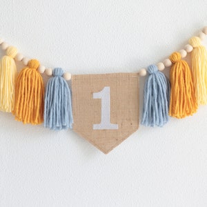 May include: A decorative banner with a burlap pennant featuring the number 1 in white. The banner is adorned with yellow, orange, and blue yarn tassels and wooden beads, perfect for a first birthday celebration.