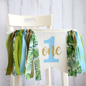 Puede incluir: Una silla alta blanca con una pancarta de tela azul y verde que dice "one" en letras doradas. La pancarta tiene cintas verdes y marrones colgando.