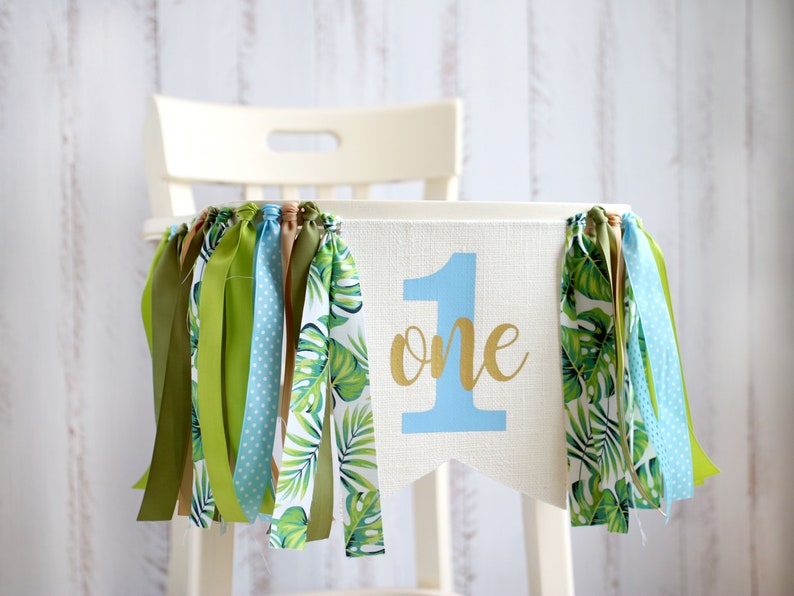 Puede incluir: Una silla alta blanca con una pancarta de tela azul y verde que dice "one" en letras doradas. La pancarta tiene cintas verdes y marrones colgando.