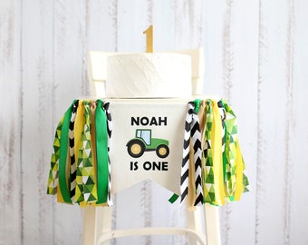 Puede incluir: Cintas de streamers verdes, amarillas y negras cuelgan de una silla alta blanca. Un pastel blanco con un adorno de oro número uno se sienta en la silla. Una pancarta verde con el texto "Noah is one" y un tractor de dibujos animados está unida a la silla.