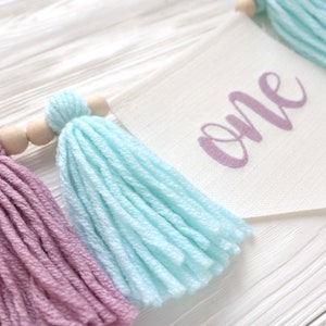 May include: A white fabric banner with the word "one" written in purple script. The banner is decorated with colorful yarn tassels in shades of pink, blue, and peach.