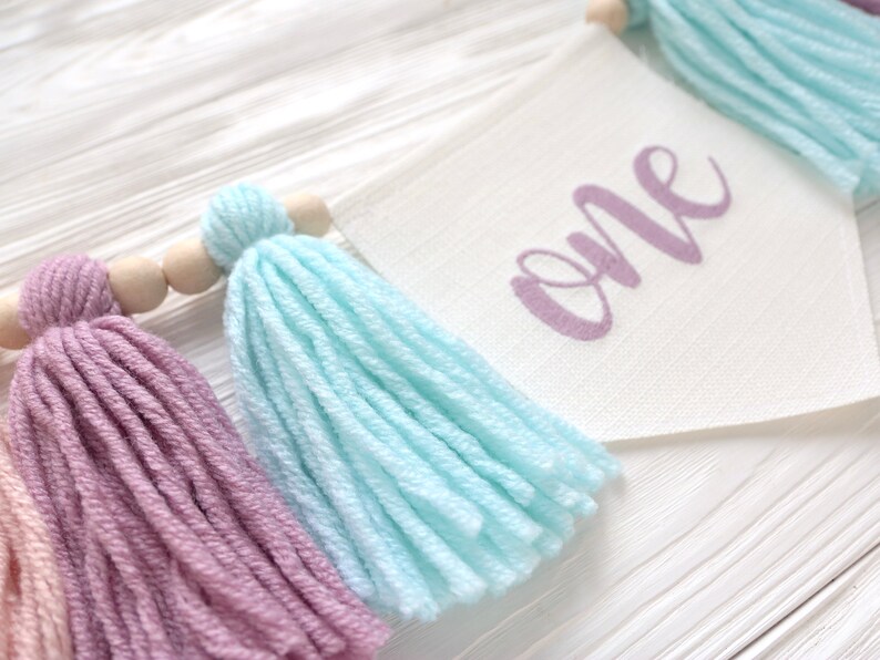 May include: A white fabric banner with the word "one" written in purple script. The banner is decorated with colorful yarn tassels in shades of pink, blue, and peach.