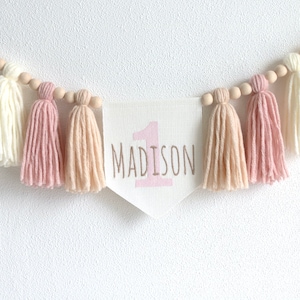 Può includere: Uno striscione bianco con frange di lana rosa e perline di legno. Lo striscione dice "1 MADISON" in lettere rosa.