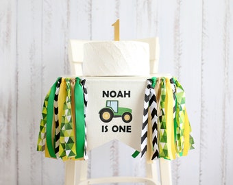 Puede incluir: Una silla alta blanca con una pancarta de tractor verde y amarillo que dice "Noah is one". La pancarta tiene cintas de colores verde, amarillo y negro colgando de ella. Un pastel blanco con un número uno dorado en la parte superior se sienta en la silla alta.