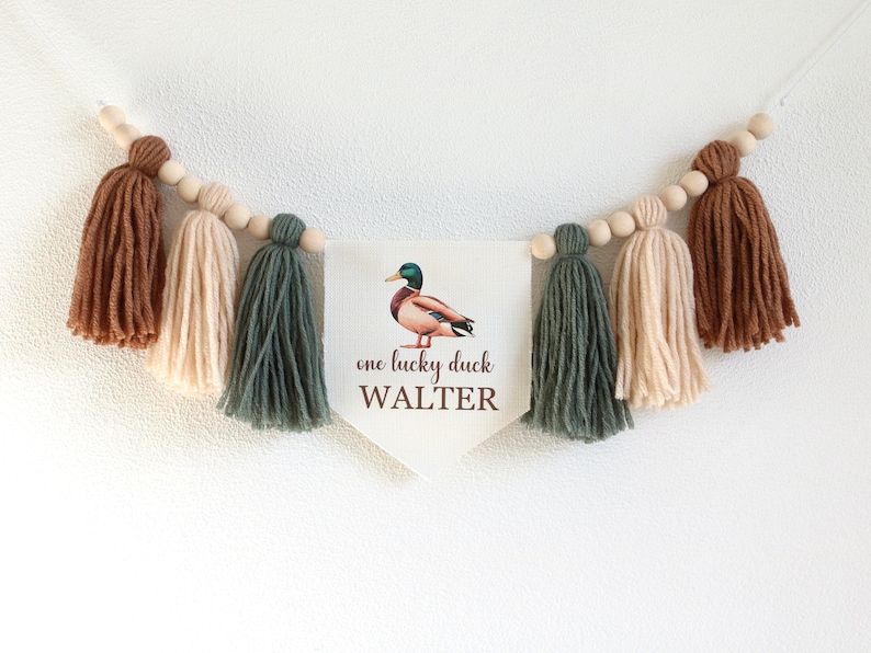 以下が含まれることがあります： 茶色のアヒルのイラストと「one lucky duck WALTER」という文字が書かれた白いバナーが、茶色、ベージュ、緑の糸のタッセルが付いた紐に吊るされています。