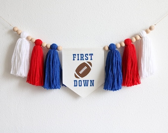 May include: A white fabric banner with the words "FIRST DOWN" in blue text and a brown football graphic. The banner is strung with a wooden bead garland with red, white, and blue yarn tassels.