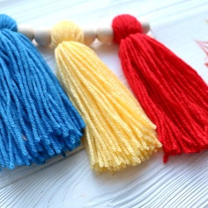 May include: Three colorful yarn tassels in blue, yellow, and red hanging from a wooden dowel. The tassels are made of soft yarn and are perfect for adding a touch of whimsy to any space.