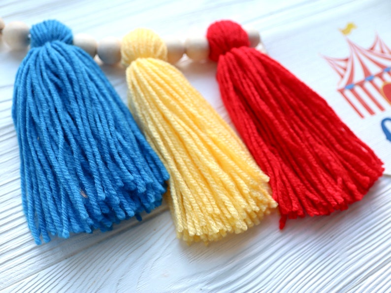 May include: Three colorful yarn tassels in blue, yellow, and red hanging from a wooden dowel. The tassels are made of soft yarn and are perfect for adding a touch of whimsy to any space.