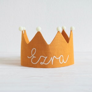 May include: A yellow linen crown with white pom poms and the name "Ezra" embroidered in white.