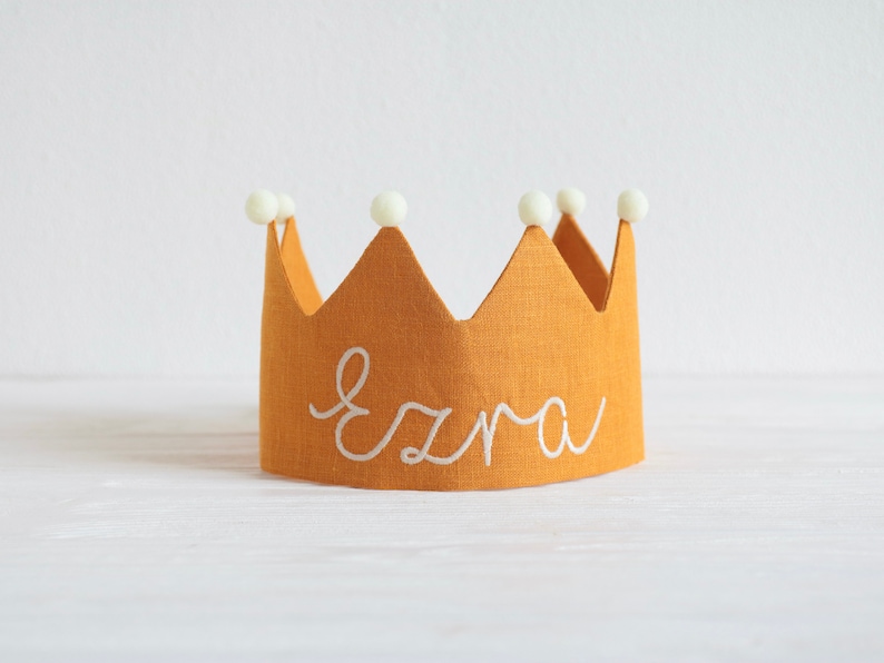 May include: A yellow linen crown with white pom poms and the name "Ezra" embroidered in white.