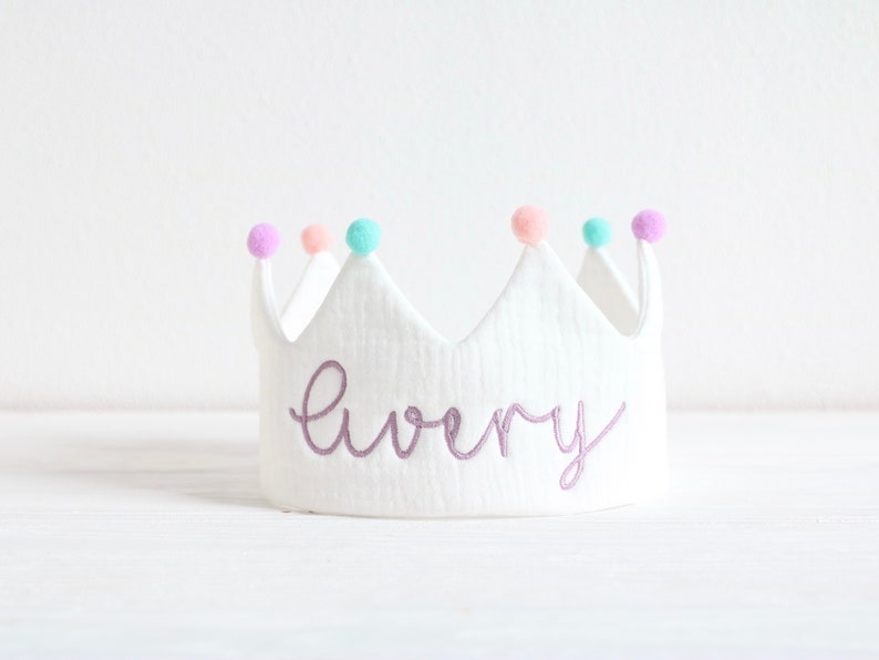 May include: White felt crown with purple pom-poms and the name "Avery" embroidered in purple thread.